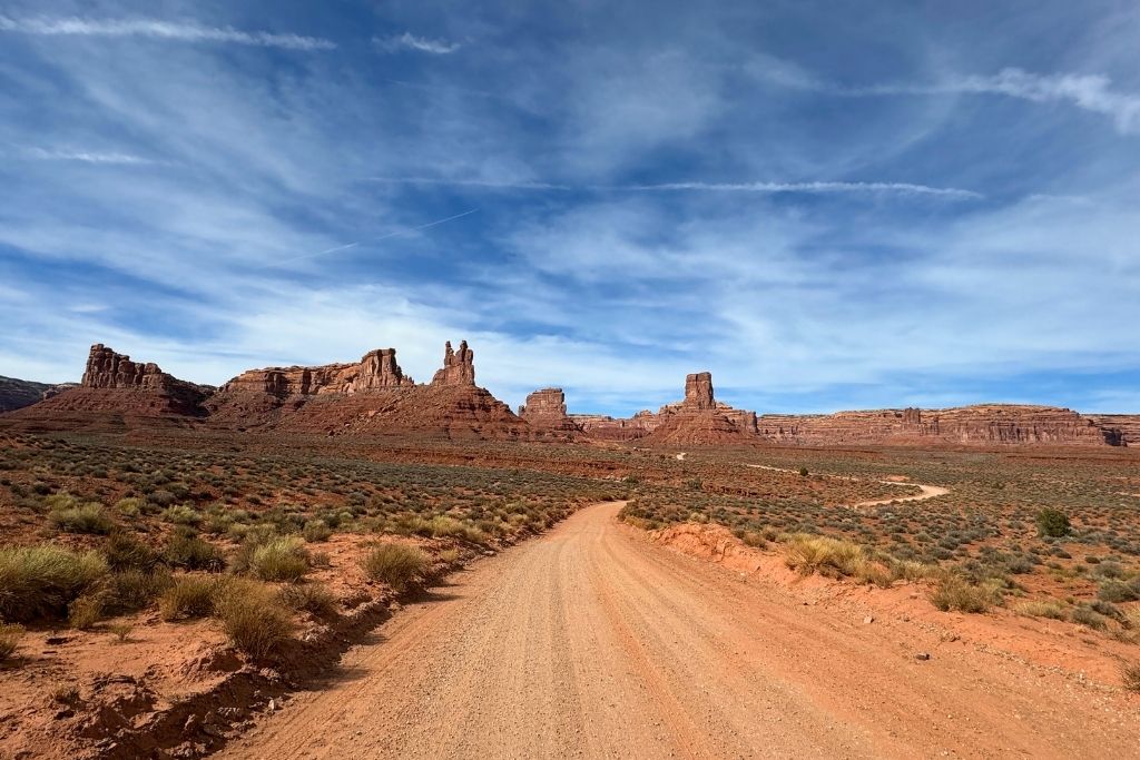 A winding dirt road cuts through the desert landscape toward towering red rock formations under a partly cloudy blue sky, capturing the scenic beauty of a road trip to Page, Arizona.