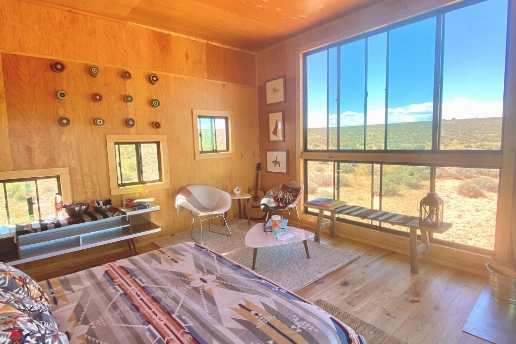 Cozy desert cabin interior with large windows overlooking the Arizona landscape, featuring wooden walls, a patterned bedspread, and eclectic decor including framed art and colorful wall accents.