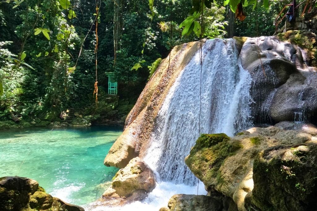 A waterfall flows down smooth rocks into a clear turquoise pool in the Jamaican jungle, surrounded by hanging vines and dense greenery.