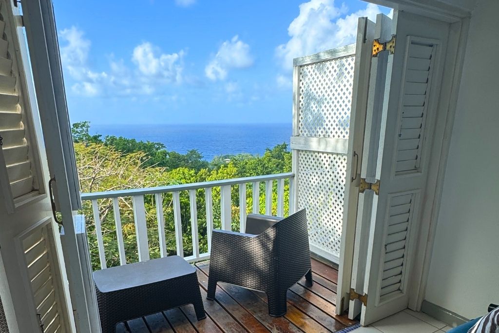 A private balcony with two modern black chairs and a small table overlooks lush greenery and the Caribbean Sea, seen through open white shutters in Jamaica.
