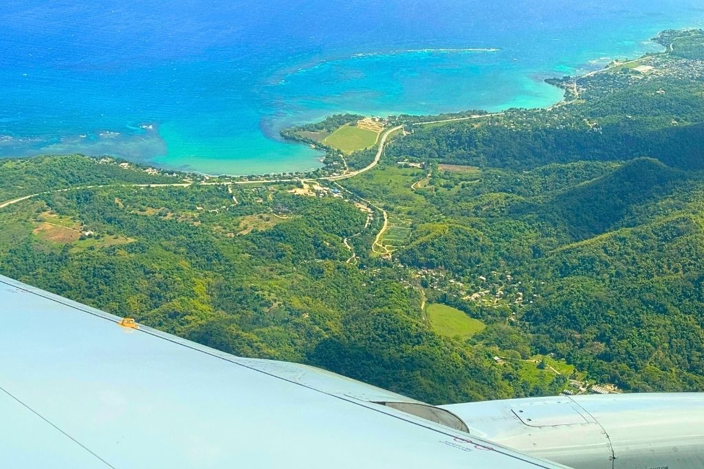 Aerial view of Jamaica’s lush green hills and turquoise coastline seen from an airplane window, with the wing visible in the foreground.