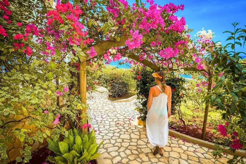 Kate walks through a stone garden path under an archway of bright pink bougainvillea flowers, dressed in a white sundress with ocean views ahead.