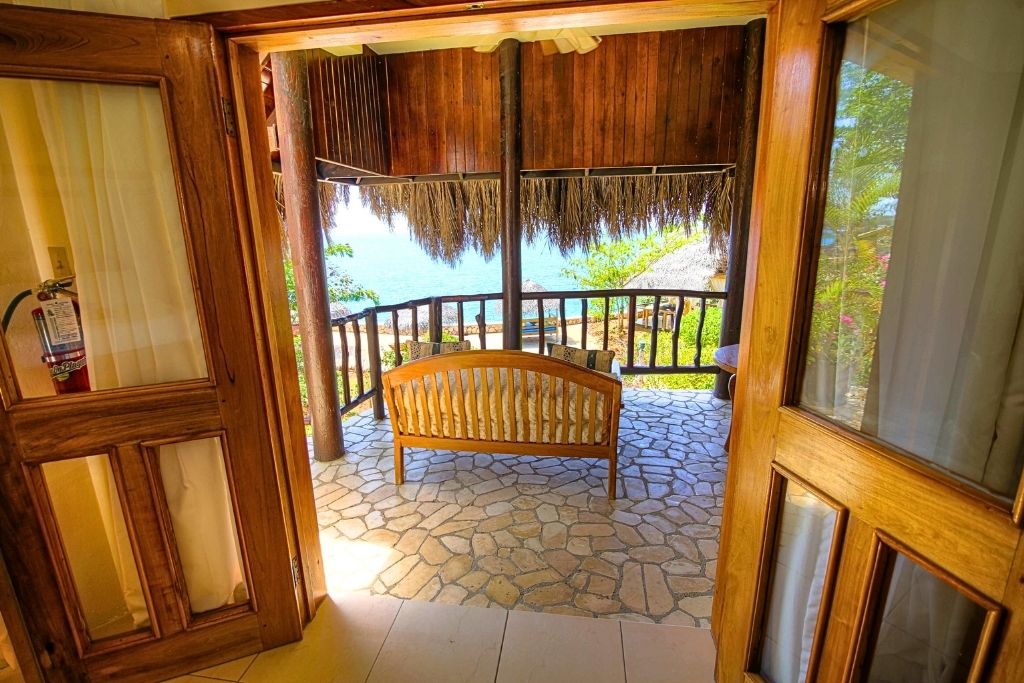 A view from inside a Jamaican resort room looking out onto a private balcony with a wooden bench, thatched roof, and ocean scenery beyond.