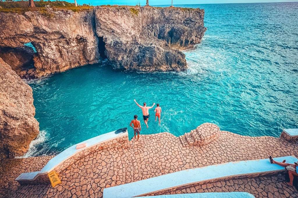 Two people leap into bright turquoise water from a stone terrace at Rick’s Cafe in Negril, Jamaica, with dramatic cliffs and ocean views all around.