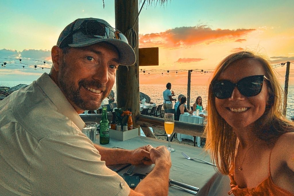 Kate and her husband smile at a seaside restaurant table in Jamaica during sunset, with drinks, string lights, and a glowing ocean backdrop behind them.