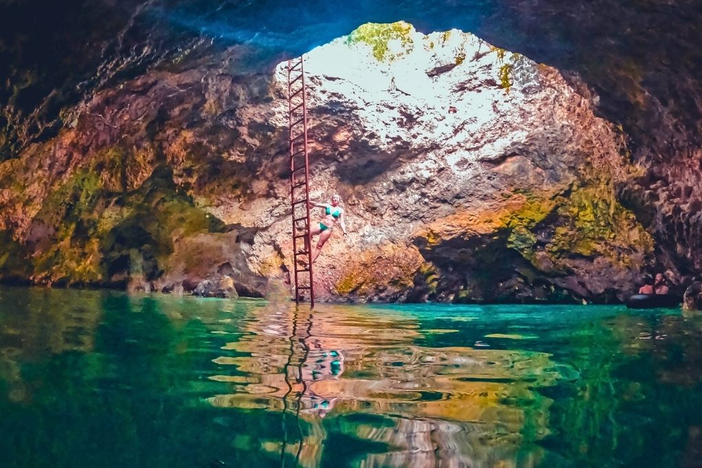 Kate climbs a tall red ladder inside the glowing Blue Hole Mineral Spring cave in Jamaica, surrounded by rocky walls and vibrant turquoise water.