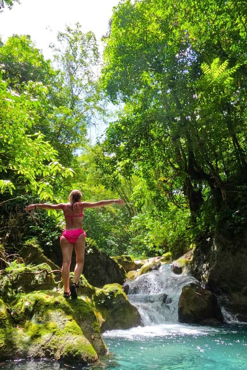 Kate stands with arms outstretched on mossy rocks above a cascading waterfall and emerald pool in the lush Jamaican jungle, wearing a pink bikini.