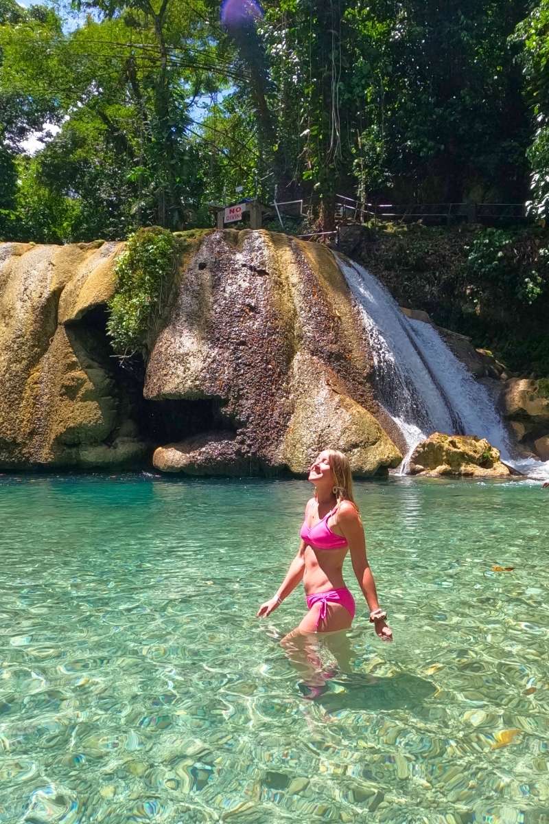 Kate stands in clear turquoise water wearing a pink bikini, smiling up toward the sun in front of a cascading jungle waterfall in Jamaica.