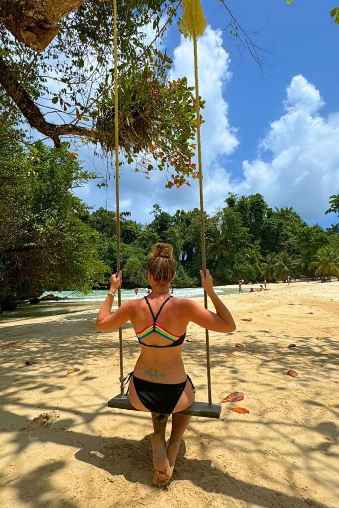 Kate sits on a rope swing facing the ocean, wearing a black bikini with a rainbow-striped top, surrounded by soft sand, lush trees, and a bright blue sky on a tropical Jamaican beach.