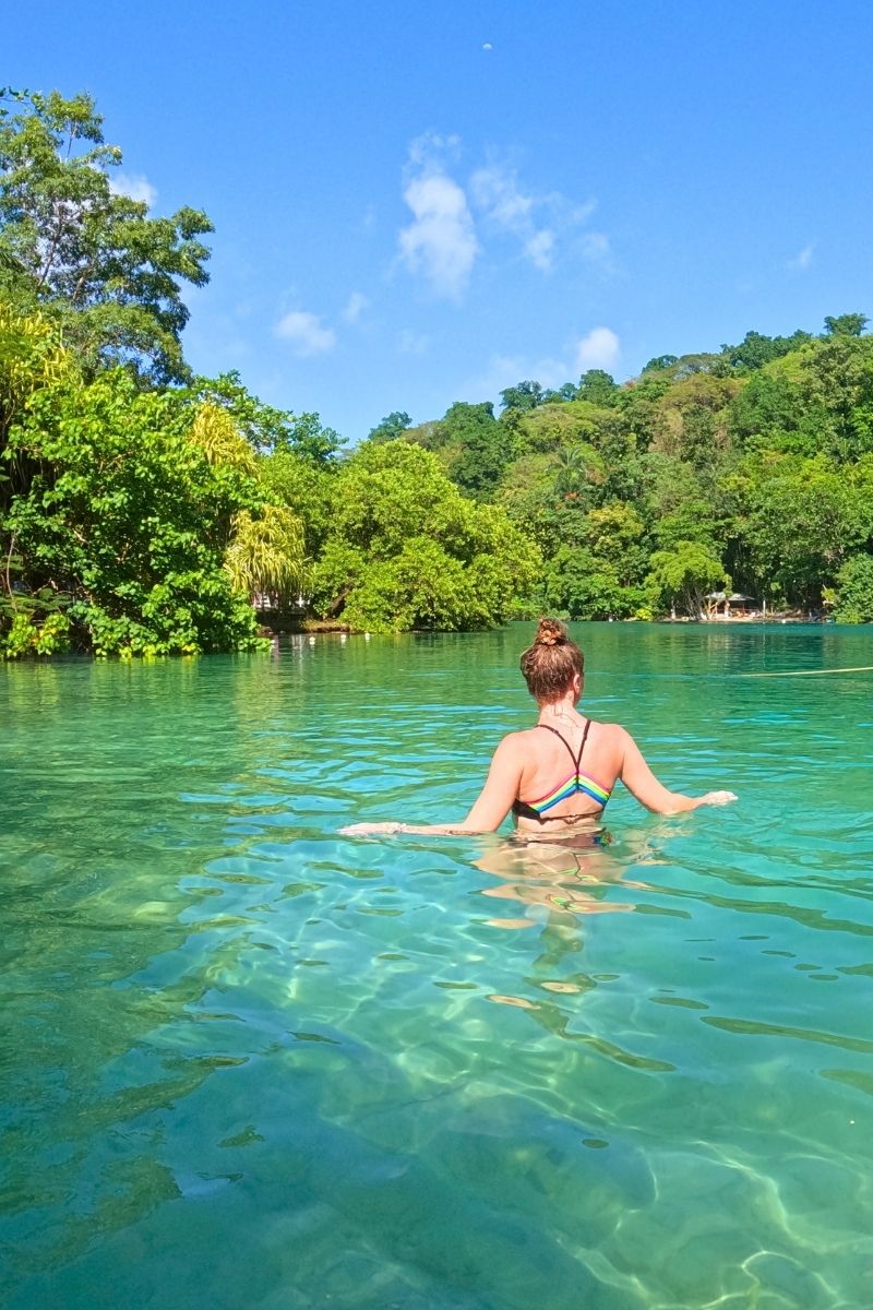A woman in a black bikini with rainbow straps wades through crystal-clear turquoise water surrounded by lush jungle in Jamaica, under a bright blue sky.
