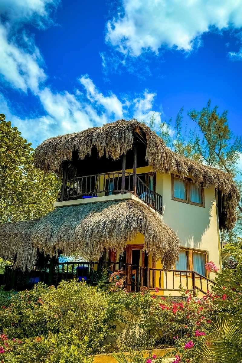 Charming two-story villa in Jamaica with thatched roofs and a wraparound porch, surrounded by lush greenery and vibrant flowers under a bright blue sky with scattered clouds.