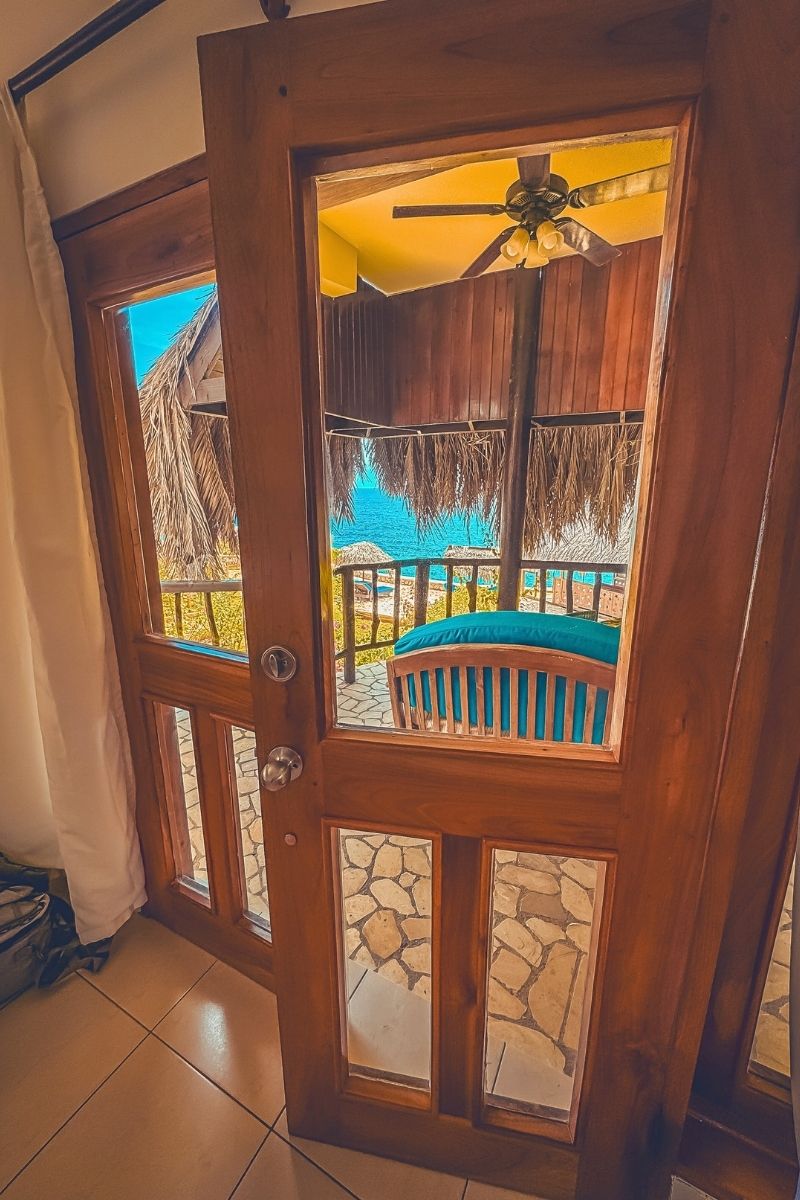 View through a wooden door with glass panels looking out onto a tropical balcony with thatched roofing, a ceiling fan, cushioned bench, and a stunning ocean view in the background.