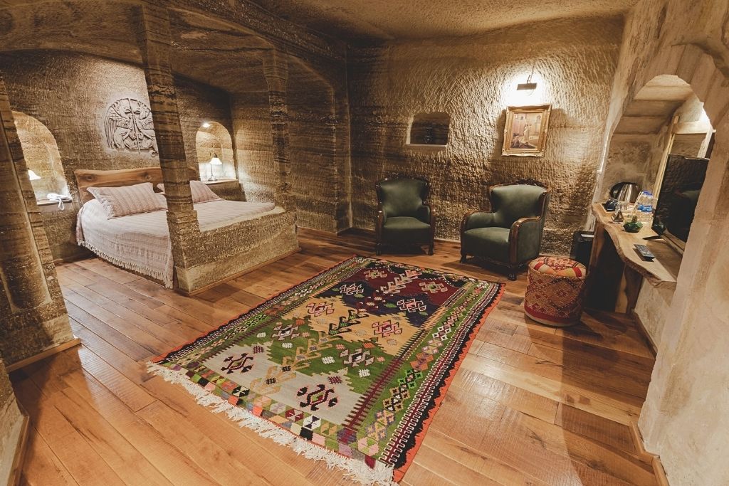 A spacious cave room at Sultan Cave Suites in Cappadocia features carved stone walls, a cozy alcove bed, colorful Turkish rug, and vintage-style seating with a wooden floor.