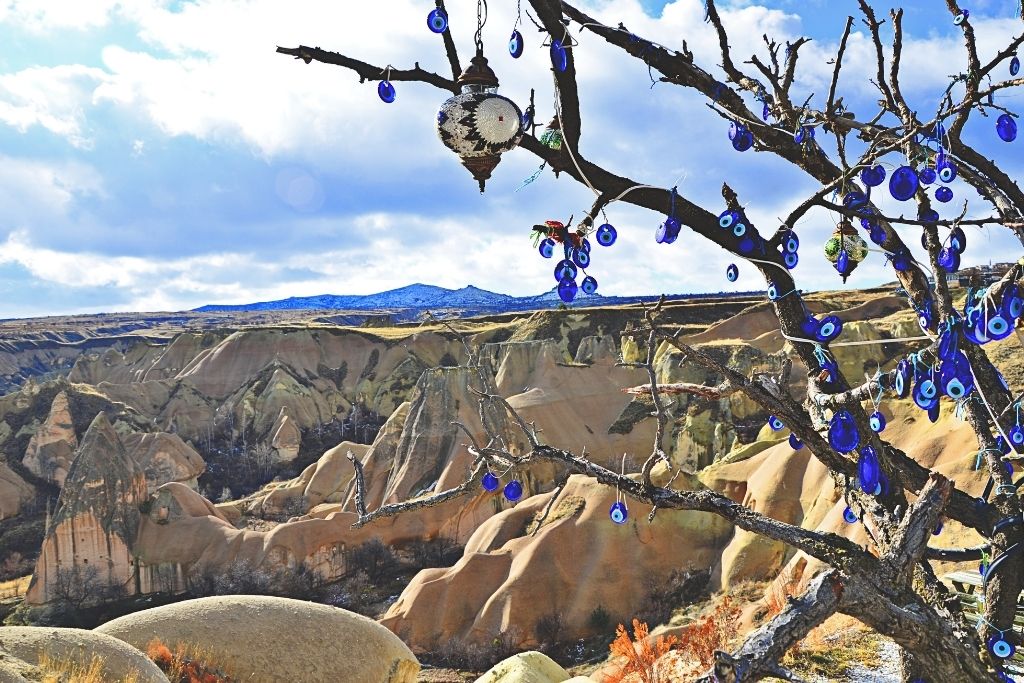 A tree decorated with vibrant blue Nazar (evil eye) charms stands at the edge of a dramatic Cappadocia valley, with soft rock formations and cloudy skies in the background.