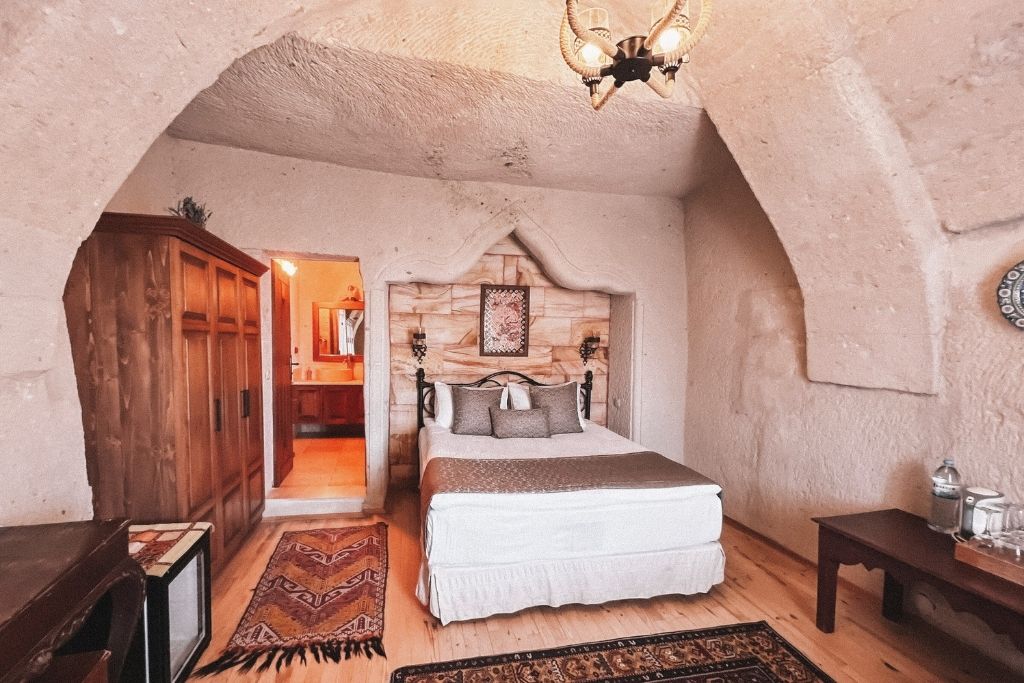 A warm-toned cave hotel room in Cappadocia featuring a carved stone arch, wooden furniture, traditional rugs, and a neatly made bed with neutral bedding.
