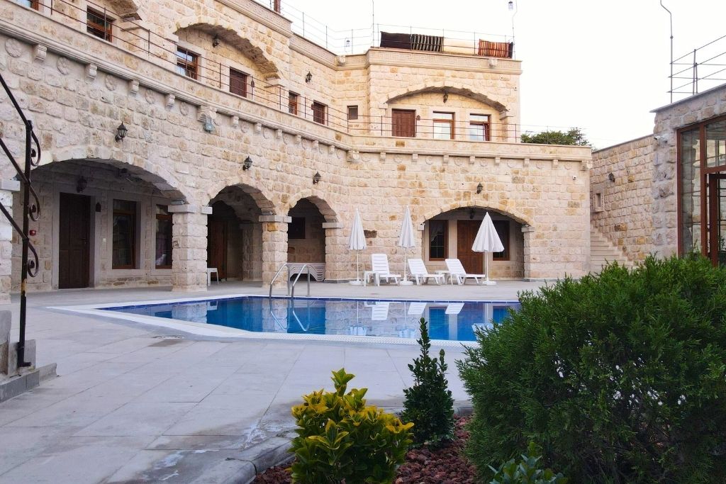 A serene courtyard pool at Göreme Cave Lodge in Cappadocia, surrounded by stone archways, white lounge chairs with umbrellas, and neatly trimmed greenery.