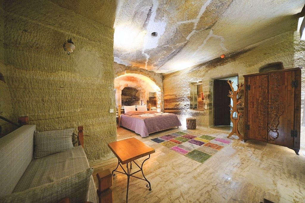 A spacious, warmly lit cave room in Cappadocia featuring textured stone walls, a cozy double bed with a purple cover, a colorful patchwork rug, and ornate wooden furniture.