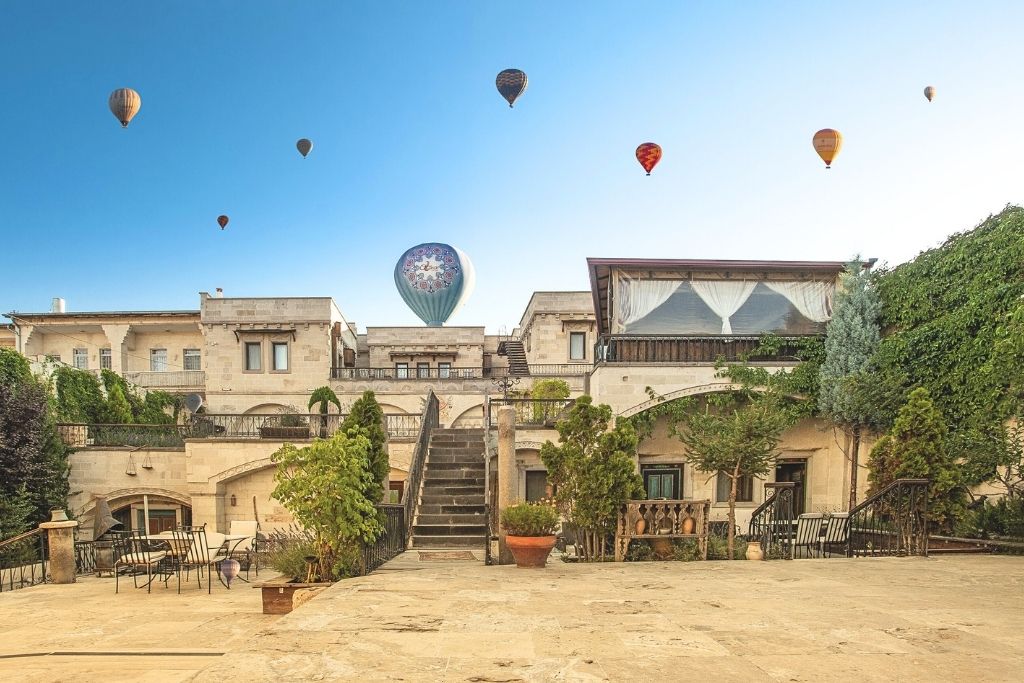 Hot air balloons rise behind the stone courtyard and lush greenery of Ottoman Cave Suites in Cappadocia, with elegant architecture and clear blue skies overhead.