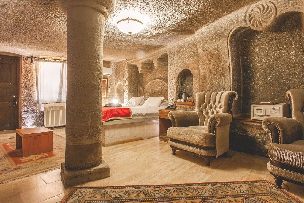 A richly textured suite at Ottoman Cave Suites in Cappadocia, featuring carved stone walls, plush armchairs, and a bed framed by traditional decor and ambient lighting.