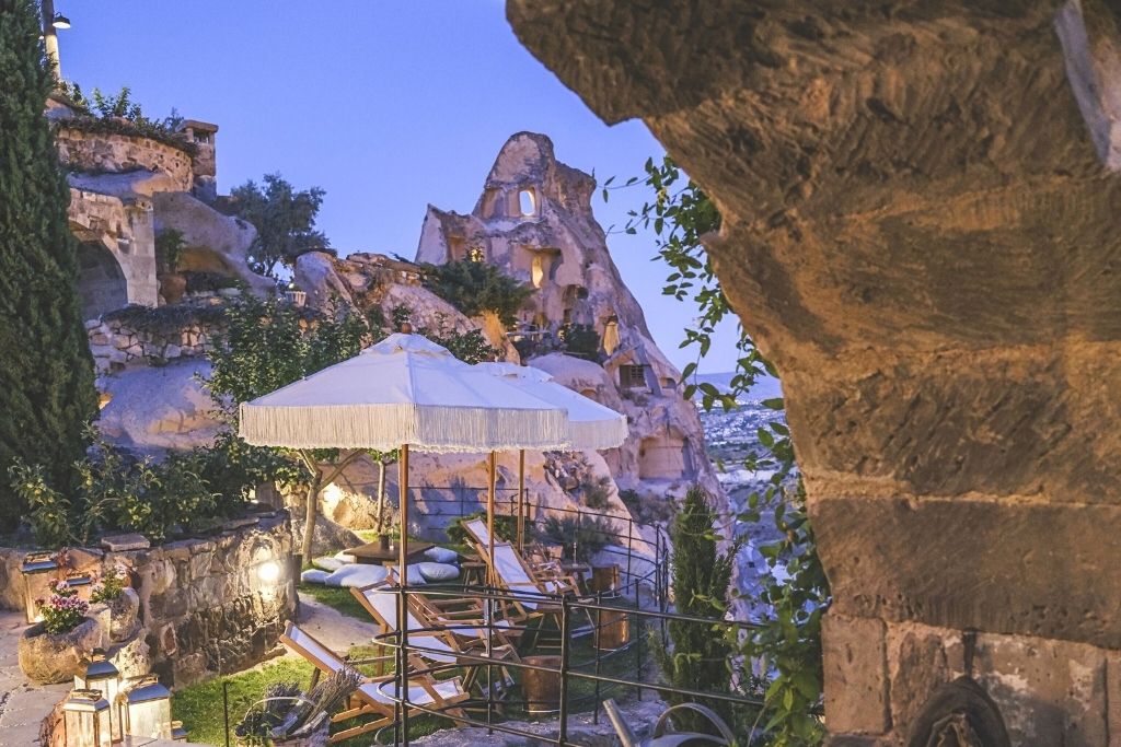A cozy terrace at Argos in Cappadocia is softly lit at dusk, with lounge chairs under a fringed umbrella set against dramatic cave dwellings and rocky cliffs.