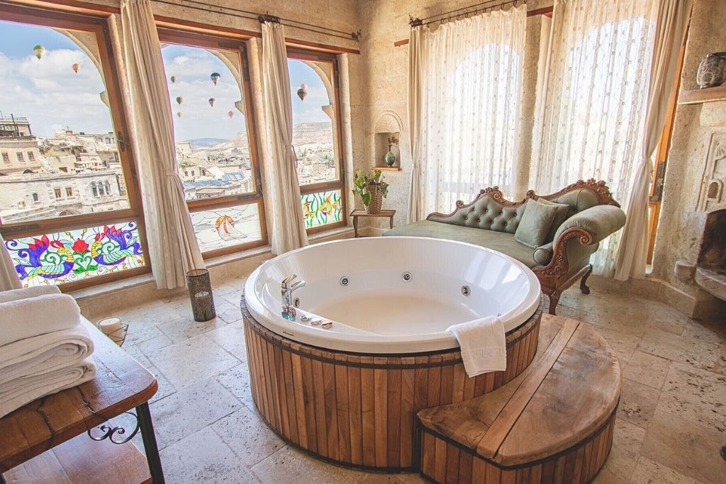 A round jacuzzi tub sits in a bright, stone-walled room at Sultan Cave Suites in Cappadocia, with vintage-style furniture and sweeping views of the town and hot air balloons outside arched windows.