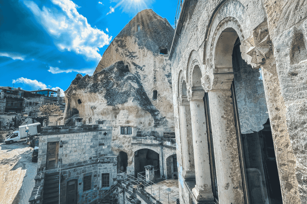 Stone archways frame a view of cave dwellings carved into soft rock under a bright blue sky in Cappadocia, with sunlight peeking over a rugged peak.