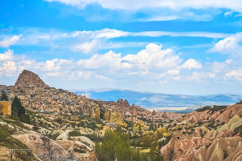 A wide view of Cappadocia’s Uchisar village nestled among pink-hued rock formations, with a bright blue sky and distant mountains in the background.