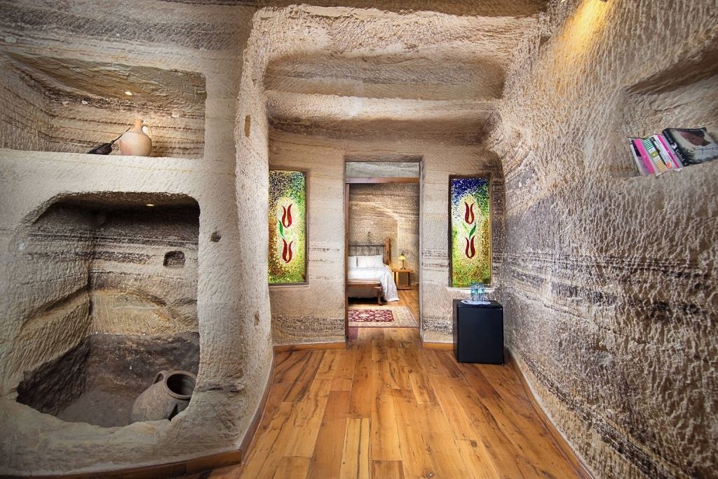 Our stylish Terra Cave Hotel suite in Cappadocia with carved stone walls, wooden floors, tulip-patterned stained glass panels, and a cozy bedroom visible through a doorway.