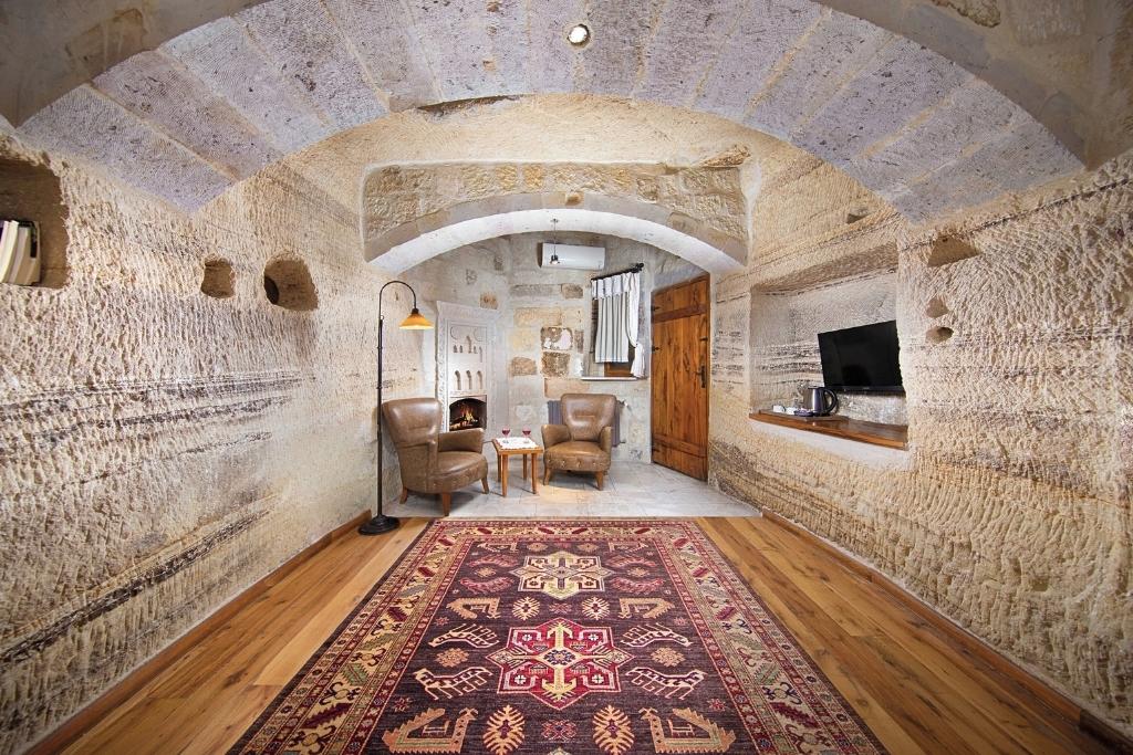Our charming Terra Cave Hotel room in Cappadocia features textured stone walls, arched ceilings, a Turkish rug, and a cozy sitting area with leather chairs and a small fireplace.
