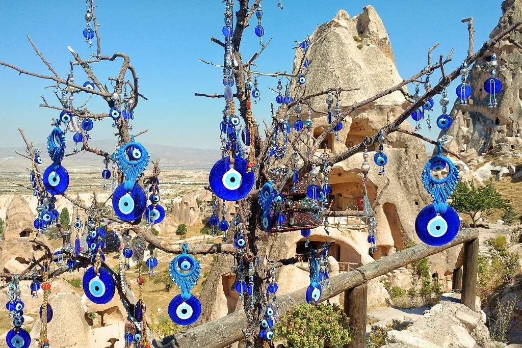A tree decorated with dozens of blue Nazar amulets (evil eye charms) stands in front of Cappadocia’s ancient cave dwellings under a clear blue sky.