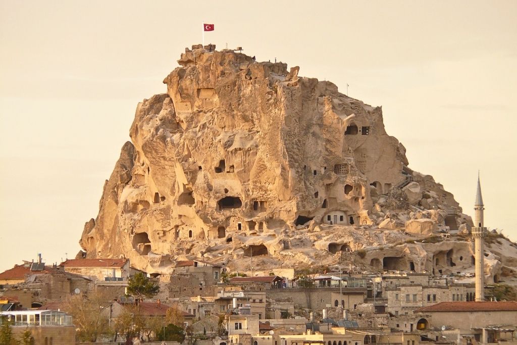 The towering rock citadel of Uchisar Castle, dotted with carved cave dwellings and topped by a Turkish flag, rises above the stone buildings of the village below at golden hour.