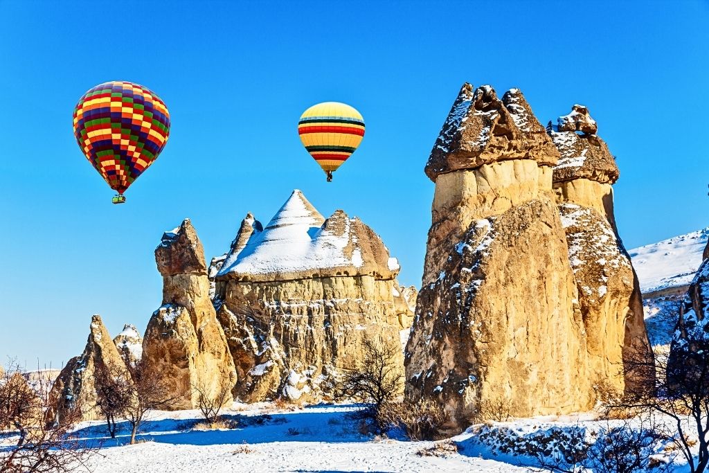 Two colorful hot air balloons float above Cappadocia’s snow-dusted fairy chimneys under a vivid blue sky on a clear winter day