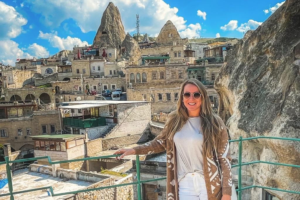 Kate poses on a rooftop terrace in Cappadocia with stone cave hotels and pointed rock formations rising behind her under a bright blue sky.