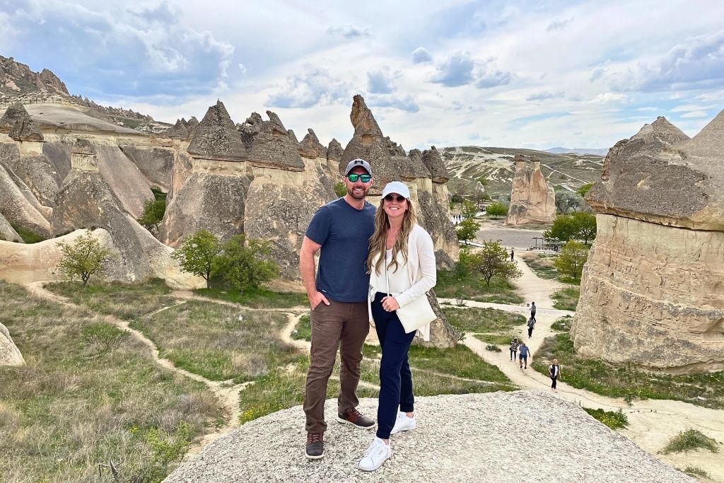 Kate and her husband smile atop a rocky ledge in Cappadocia, surrounded by the region’s iconic fairy chimneys and scenic hiking trails.