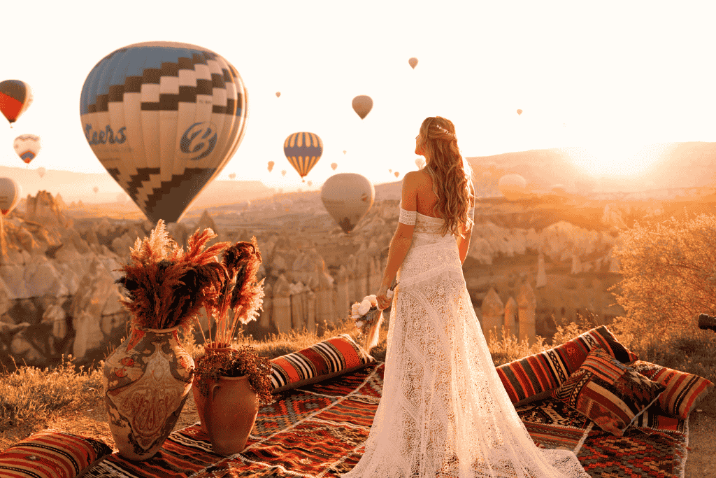 Kate stands in a white lace dress on a carpeted lookout at sunrise in Cappadocia, surrounded by hot air balloons floating over the rocky landscape.