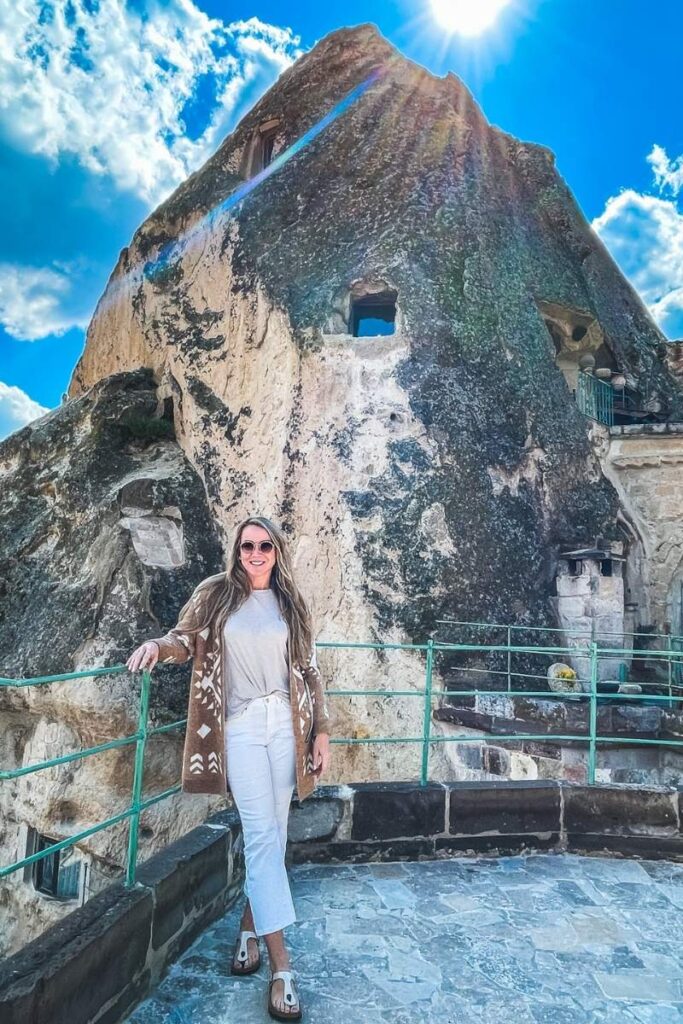 Kate poses on a stone terrace in front of a cave-carved rock formation in Cappadocia, smiling beneath a bright sunburst and vivid blue sky.