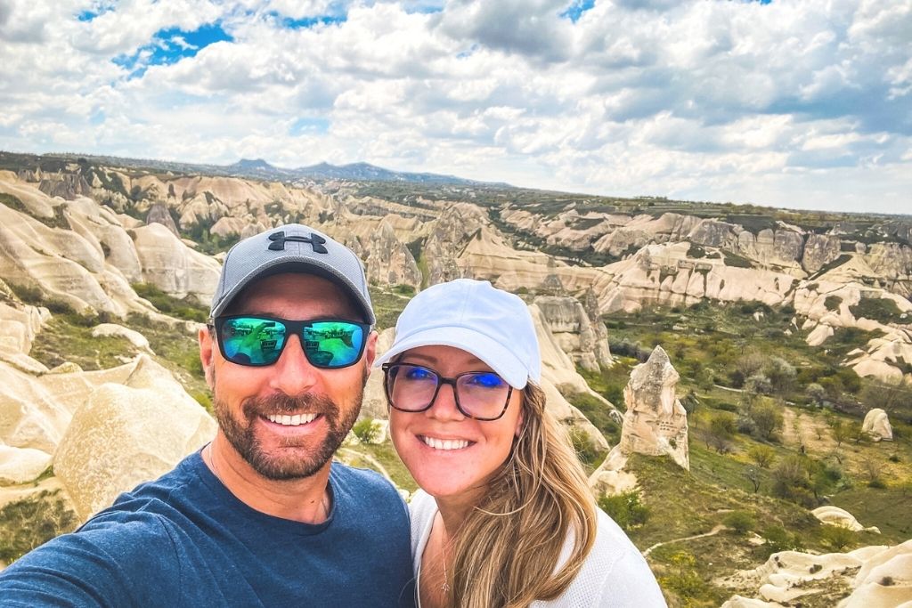 Kate and her husband smile for a selfie in Pigeon Valley, with Cappadocia’s distinctive rock formations and sweeping landscape behind them under a partly cloudy sky.