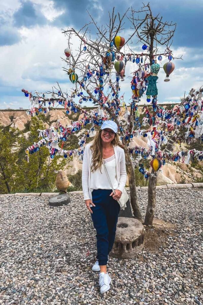 Kate stands in front of an evil eye tree in Cappadocia, decorated with Nazar charms, hot air balloon ornaments, and colorful ribbons against a rocky valley backdrop.