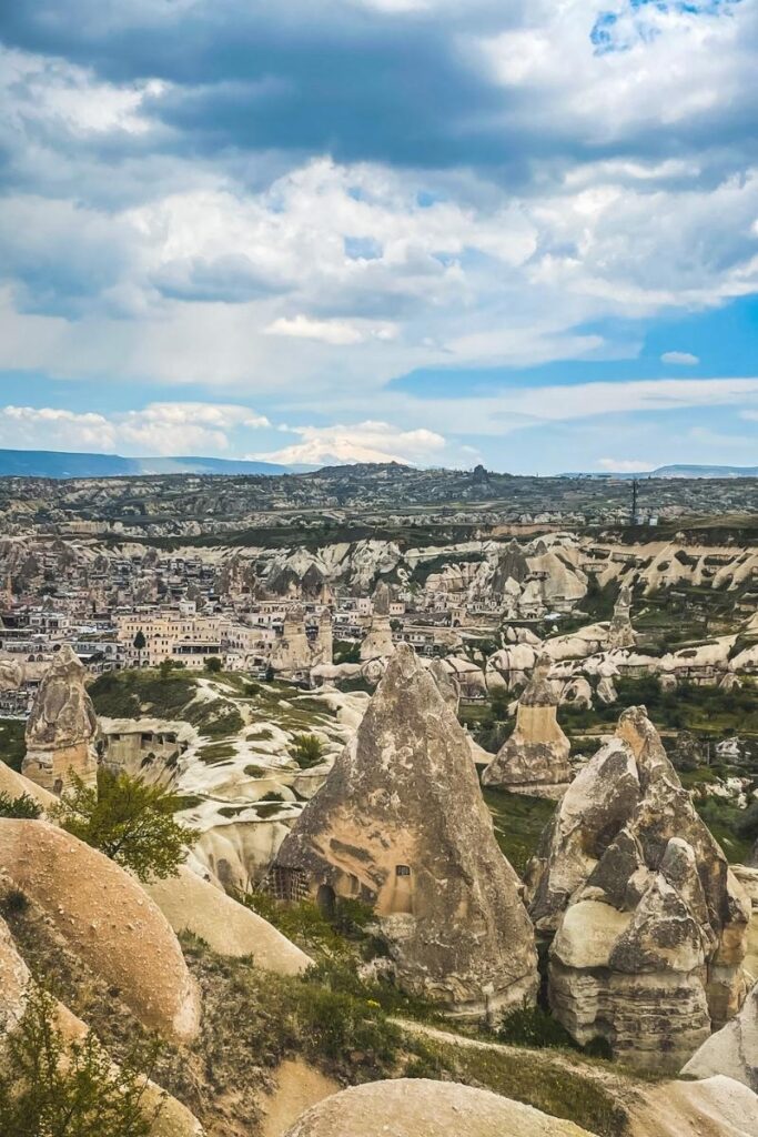 A panoramic view of Pigeon Valley in Cappadocia, showing pointed rock formations and a village nestled among the soft, sculpted landscape under a dramatic sky.