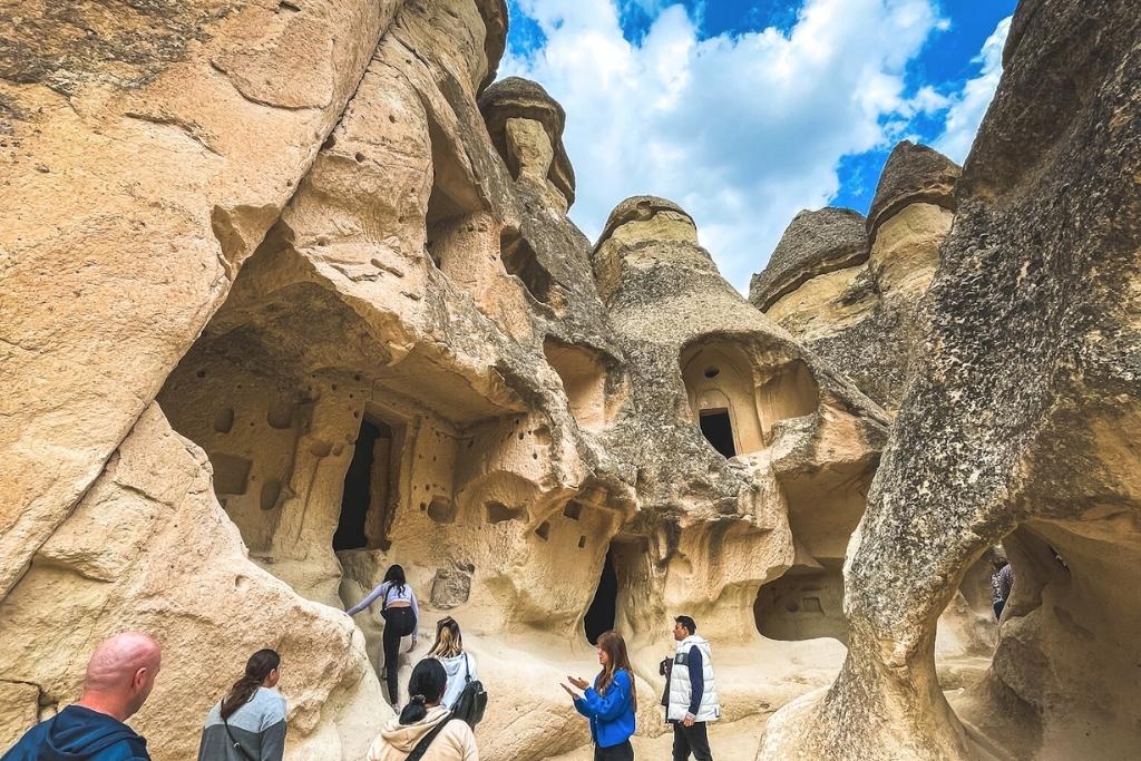 Visitors explore the carved rock dwellings of Monks Valley in Cappadocia, walking through honeycombed fairy chimneys under a partly cloudy sky.