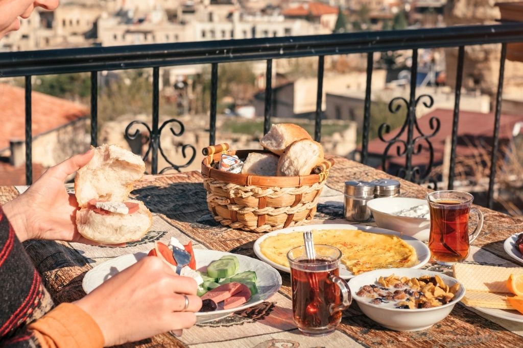 A traditional Turkish breakfast is served on a sunlit terrace in Cappadocia, featuring bread, omelet, sliced vegetables, tea, yogurt, and cereal with a view of the town below.