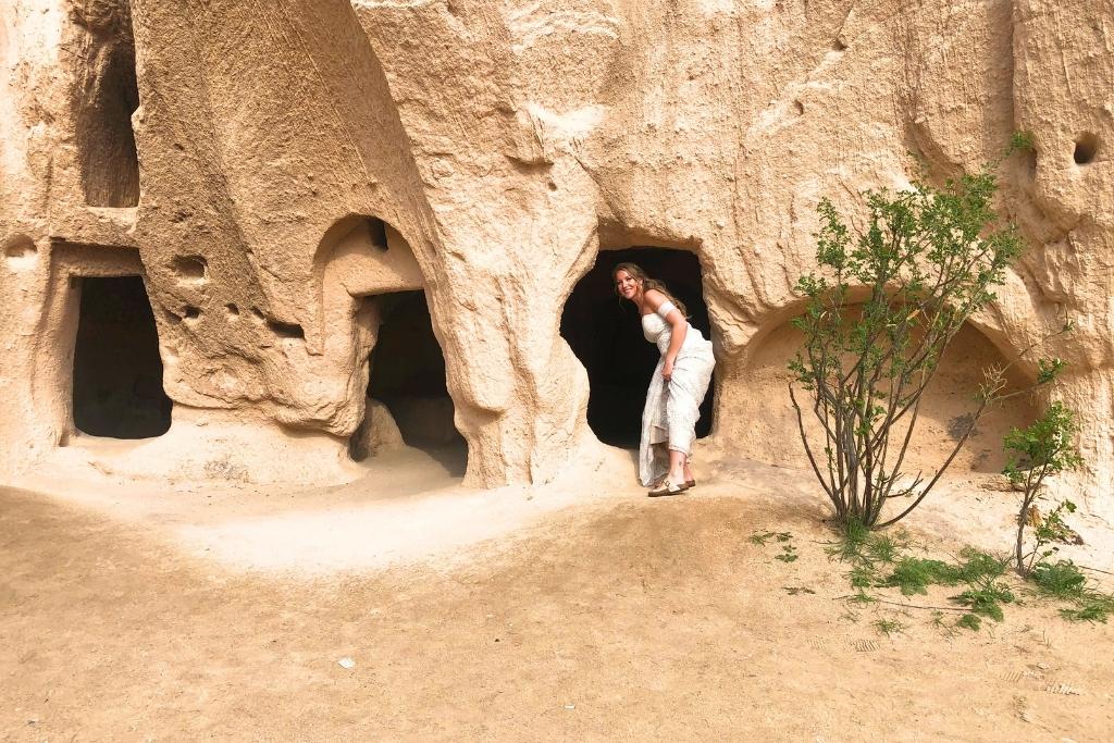 Kate, dressed in a white lace dress, bends slightly to enter a carved cave doorway in Cappadocia, surrounded by smooth rock walls and sparse greenery.