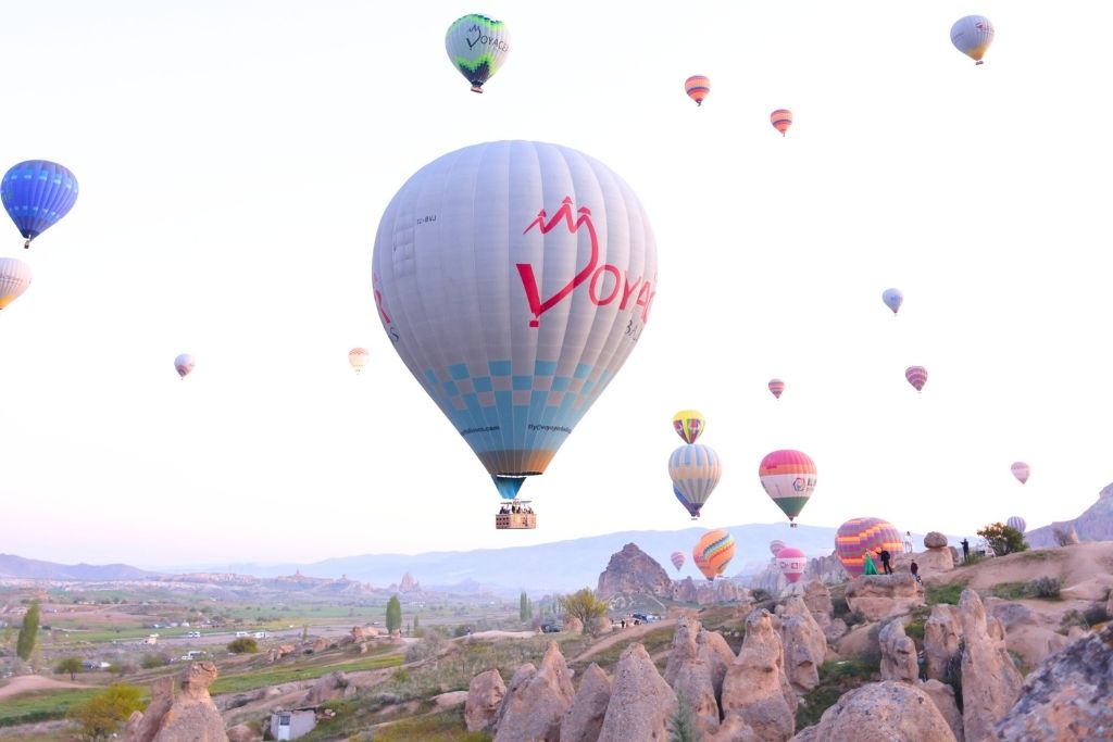 Colorful hot air balloons float above the rocky landscape of Cappadocia at sunrise, with the "Voyager" balloon prominently in the foreground against a pale sky.