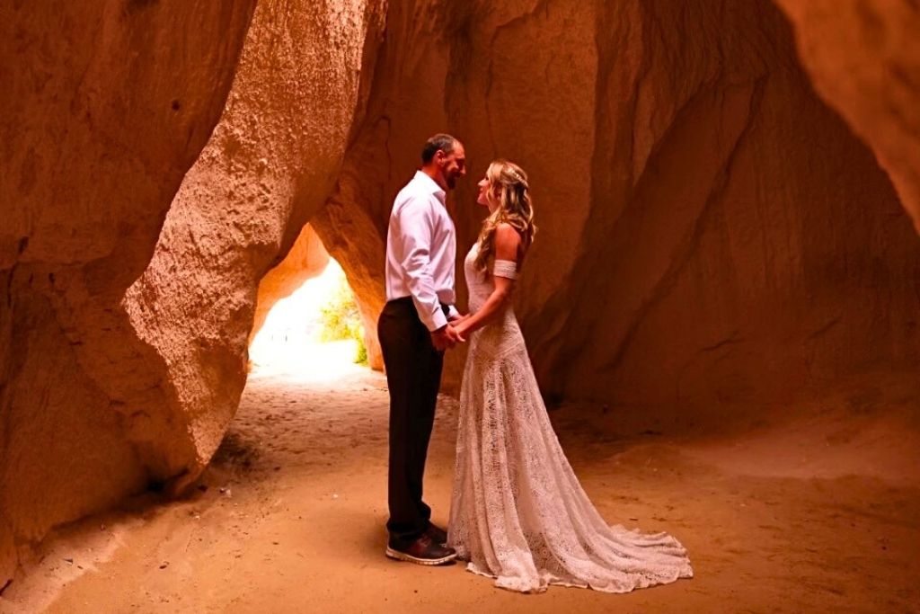 Kate and her husband stand hand in hand inside a sunlit cave in Cappadocia, her lace wedding dress trailing behind her as they share a quiet, romantic moment.