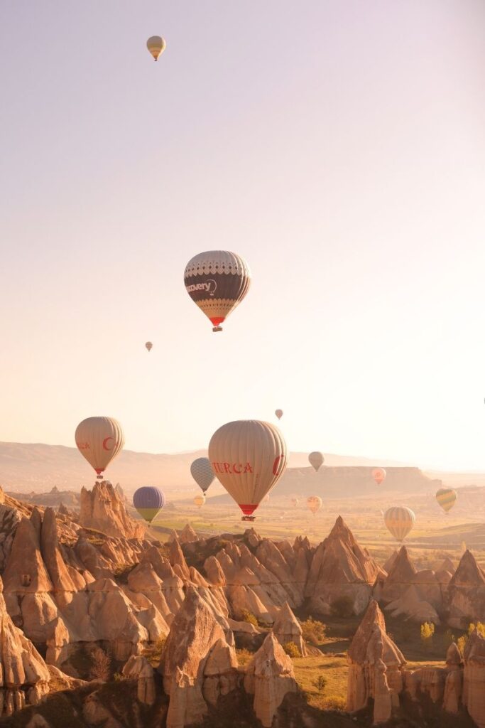 Hot air balloons drift over the jagged rock formations of Cappadocia at sunrise, casting soft shadows across the glowing valley below.
