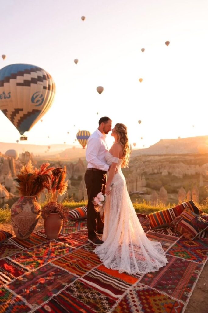 Kate and her husband share an intimate kiss at sunrise in Cappadocia, surrounded by hot air balloons and vibrant Turkish rugs on a scenic overlook.