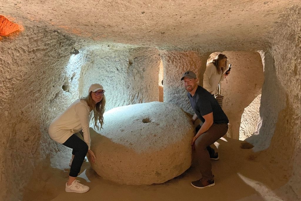 Kate and her husband crouch beside a large stone wheel door inside the Derinkuyu underground city in Cappadocia, smiling as they explore the carved tunnels.