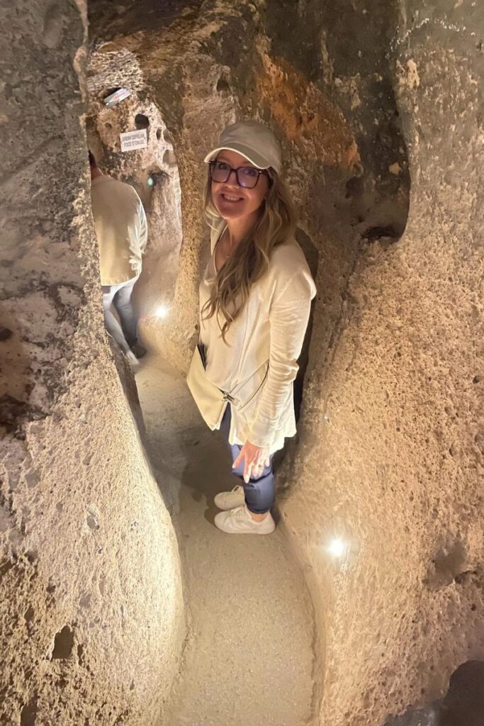 Kate smiles while walking through a narrow, dimly lit tunnel in the Derinkuyu underground city in Cappadocia, surrounded by carved volcanic rock.