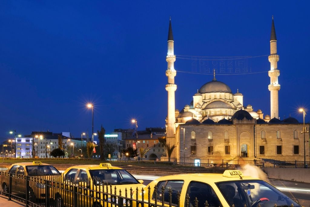 A row of yellow taxis lines the street at dusk in Istanbul, with the illuminated Yeni Cami (New Mosque) and its towering minarets glowing against the deep blue evening sky.