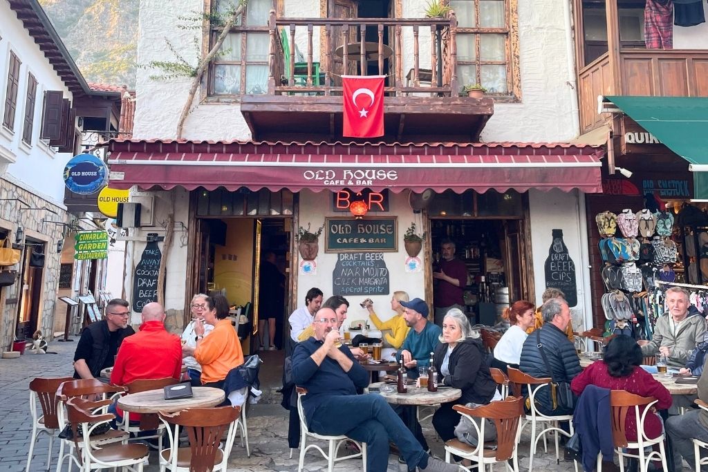 Outdoor diners gather at Old House Café & Bar in Turkey, sitting beneath a red awning and Turkish flag in a lively cobblestone alley lined with restaurants and shops.